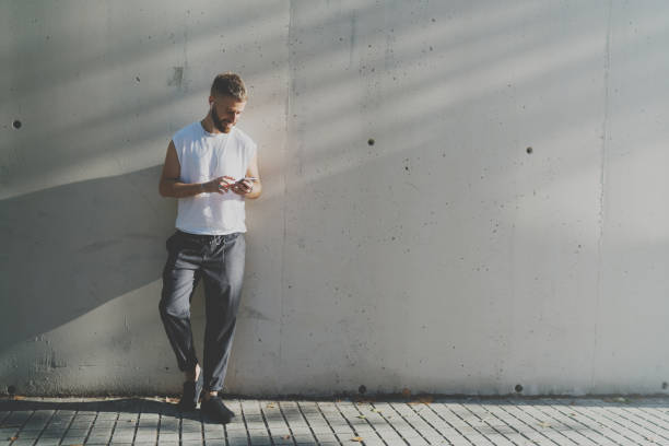 istock Successful entrepreneur watching news in internet on a mobile phone. Smiling hipster guy surfing web on a cell phone while standing against light gray concrete wall with a blank area. 1025963688