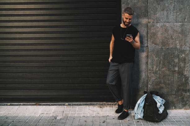 istock Smiling hipster guy surfing web on a cell phone while standing against black and gray street wall with a blank area. Young entrepreneur usinf mobile phone apps for searching the route to a hotel. 1025963788