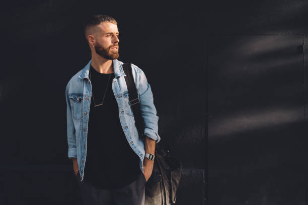 retrato de longitud completa de un hombre guapo con barba vestido con ropa de moda permanente en la calle contra la pared de hormigón rblack. chico guapo hipster posando en una chaqueta negra, camiseta y pantalones vaqueros. - chaqueta tejana fotografías e imágenes de stock