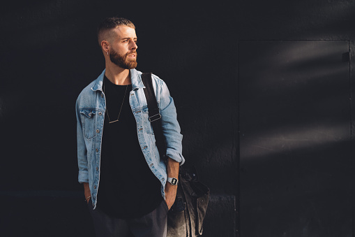 Full length portrait of a handsome bearded man dressed in fashionable clothes standing on a street against the rblack concrete wall. Handsome hipster guy posing in a black t-shirt and jeans jacket.