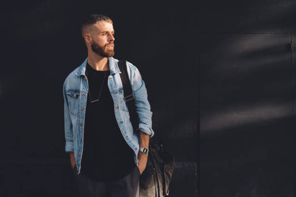 Full length portrait of a handsome bearded man dressed in fashionable clothes standing on a street against the rblack concrete wall. Handsome hipster guy posing in a black t-shirt and jeans jacket. Full length portrait of a handsome bearded man dressed in fashionable clothes standing on a street against the rblack concrete wall. Handsome hipster guy posing in a black t-shirt and jeans jacket. denim jacket stock pictures, royalty-free photos & images