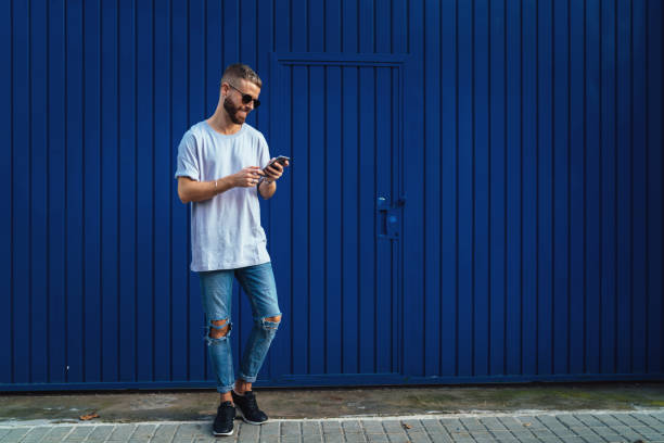 istock Smiling student guy chatting online  via social network on a mobile phone while walking the street. Stylish hipster male checking email box on a smartphone while standing on a blue wall background. 1025963798