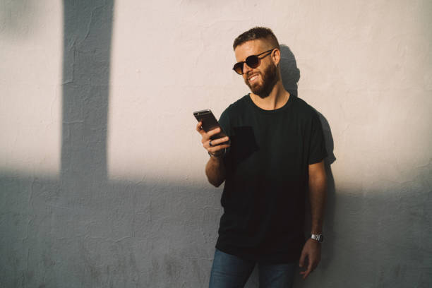 istock Smiling entrepreneur guy reading emails on a mobile phone while walking the street. Stylish hipster typing messages on a smartphone while standing on a white concrete wall background. 1025963806