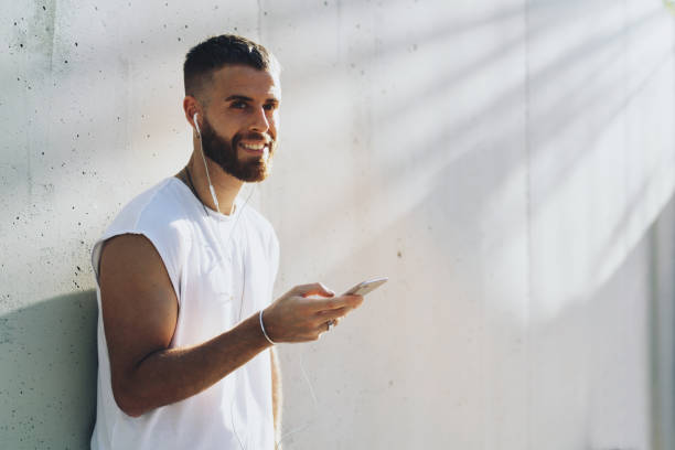 istock Smiling student guy chatting online via social network on a mobile phone while standing outdoors. Stylish hipster male checking email box on a smartphone while standing on a white wall background. 1025963844