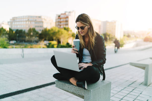 foto de mujer disfrutando de tomar café a trabajar al aire libre en un ordenador portátil conectado a wifi pública independiente. chica elegante estudiante estudiar en línea y dedicando el tiempo libre en un parque - hipster people surfing the net internet fotografías e imágenes de stock