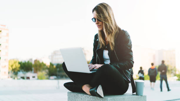 young fashion blogger wearing stylish clothes writing new post in her blog by a laptop connected to wifi while sitting in a park with take away coffee in a paper cup on a warm sunny day. - hipster people surfing the net internet imagens e fotografias de stock
