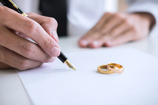 Hands of husband signing decree of divorce (dissolution or cancellation) of marriage filing divorce papers and two golden marry ring.
