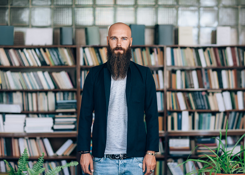 Portrait of bearded hipster man in library. Bald man with long beard stands on background of books. Education concept