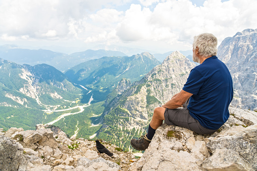 Senior man sitting on the edge of rocks and looking into the mountain range in distance. Side view.