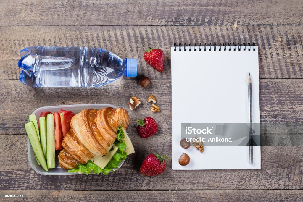 Caixa de almoço com croissants, frutas e legumes em notebook com lugar vazio para texto e fundo de madeira. Copie o espaço. Volta ao conceito de escola - Foto de stock de Almoço royalty-free