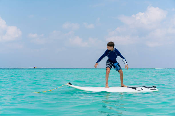 Young boy balancing on surfboard against blue sky on sandy bikini beach on Gulhi island, Maldives. Indian ocean, clouds on blue sky and horizon are in background. Gulhi island not far from Maafushi island on Maldives and is open for foreigners.