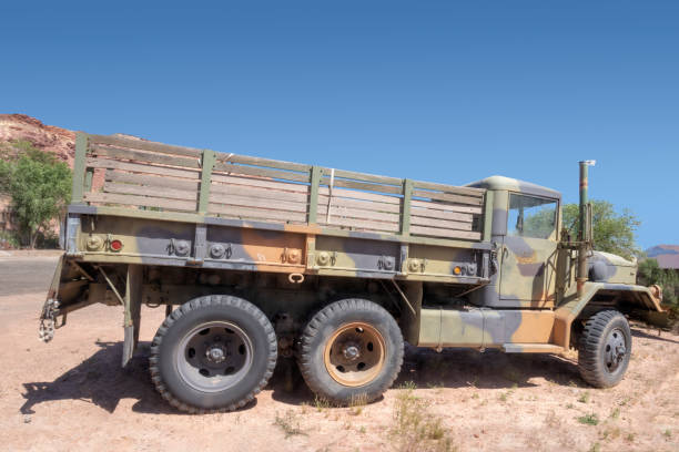 Two old military trucks under a metal shelter, dusty and overgrown with weeds, in an abandoned storage area.