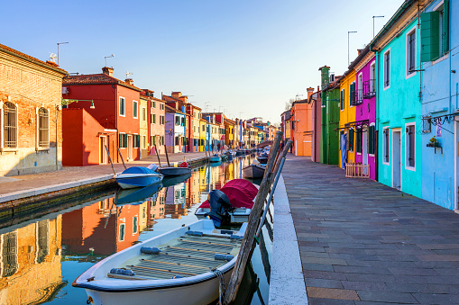 Colorful canal houses and boats on Burano island in the Venetian Lagoon. A Venice landmark in northern Italy. Vessel and buildings reflecting in calm water. Beautiful poster like travel and tourism background on a sunny day with copy space.