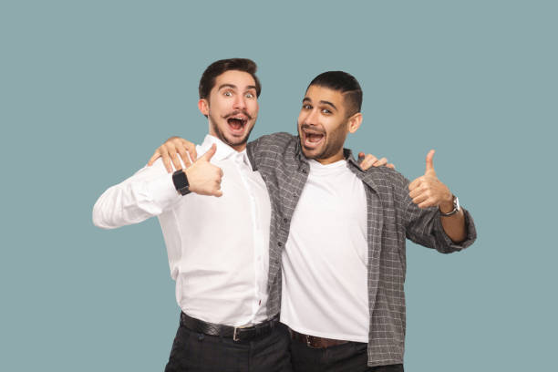 Portrait of two handsome bearded happy friends or partners standing hugging, and looking at camera with satisfied amazed face and thumbs up. Portrait of two handsome bearded happy friends or partners standing hugging, and looking at camera with satisfied amazed face and thumbs up. indoor studio shot, isolated on light blue background. young handsome turkish man portrait stock pictures, royalty-free photos & images
