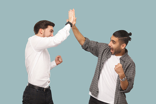Yes we did. expression profile side view of two young handsome happy satisfied bearded partner celebrating triumph together, giving hi five hands. indoor studio shot, isolated on light blue background