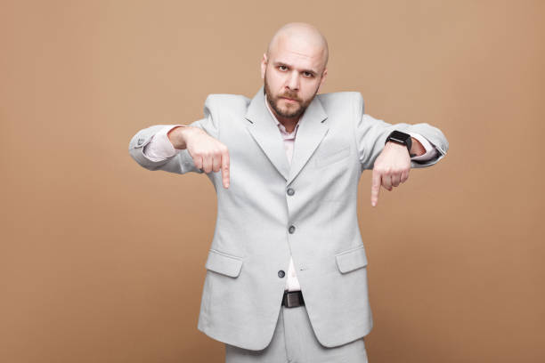 istock Here and right now. Serious middle aged bald bearded businessman boss in classic light gray suit standing, looking at camera and showing pleace. 1026202374