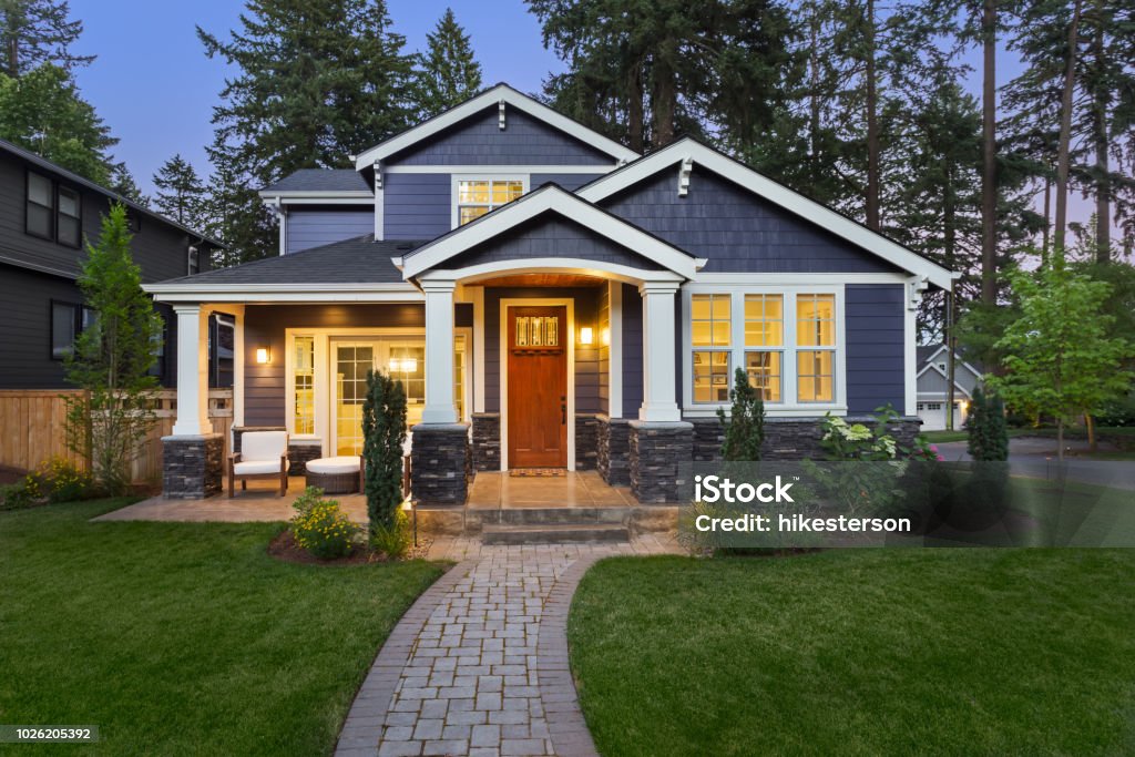 Beautiful luxury home exterior at twilight facade of home with manicured lawn, landscaping, and backdrop of trees and dark blue sky. Glowing interior lights create a welcoming mood. House Stock Photo