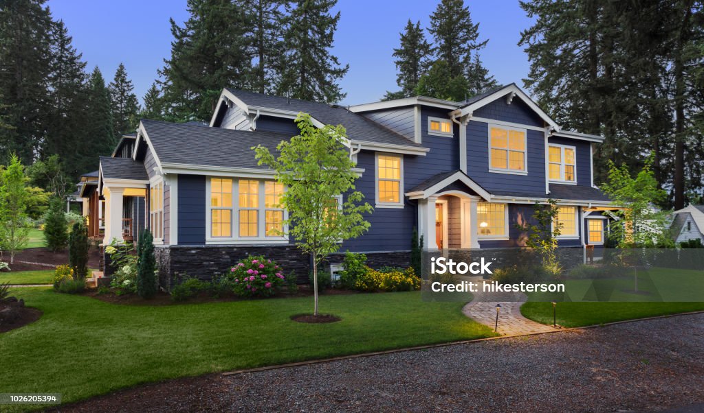 Beautiful luxury home exterior at twilight facade of home with manicured lawn, landscaping, and backdrop of trees and dark blue sky. Glowing interior lights create a welcoming mood. House Stock Photo