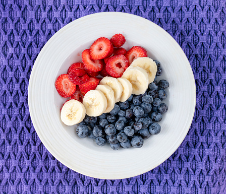 Conceptual image. A bowl of fruit from directly above. Strawberries, bananas, blueberries in white bowl on purple mat. The colors red, white, blue and purple represent symbolic political USA colors: red strawberries Republicans, blue blueberries Democrats, dividing white banana line and purple mat for a combination of red and blue.