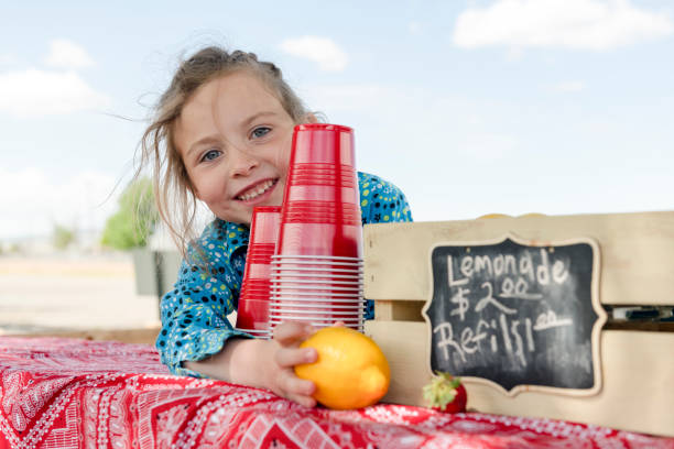 Little girl selling drinks at lemonade stand A little girl selling lemonade reaches for a lemon and smiles. Sign indicates price of drinks. images of lemonade stands stock pictures, royalty-free photos & images