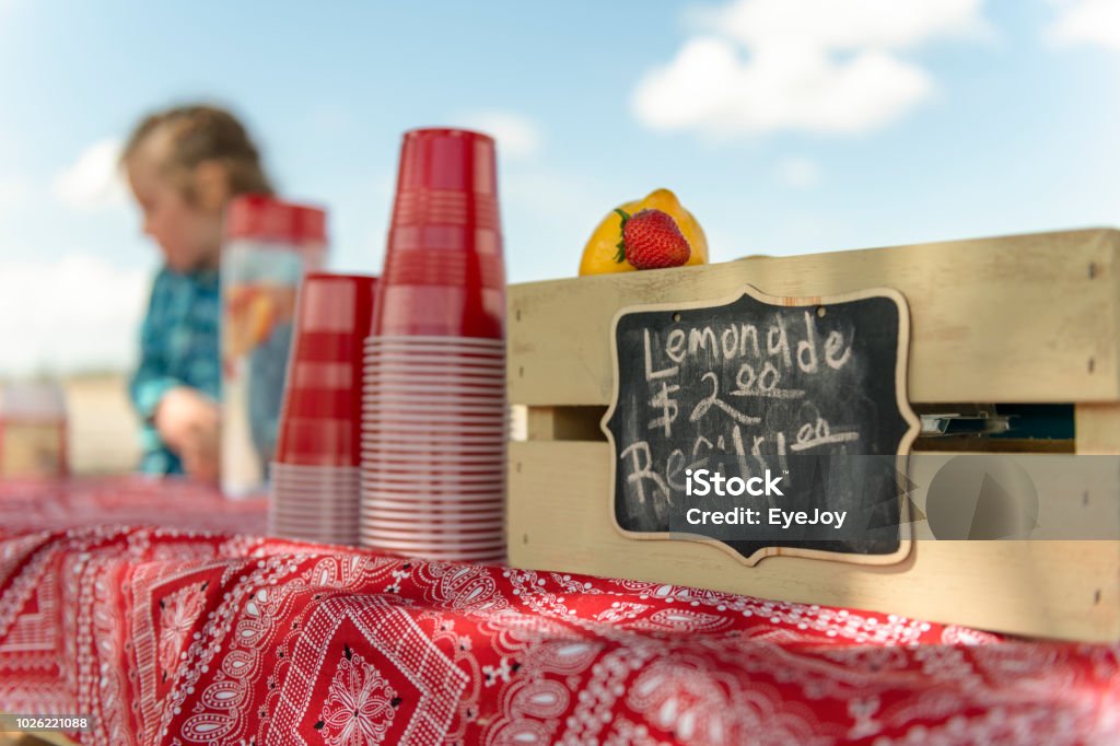 Lemonade stand and sign Focus on sign at lemonade stand. A lemon, strawberry and pile of red plastic cups in foreground. Out of focus girl in background. Child Stock Photo