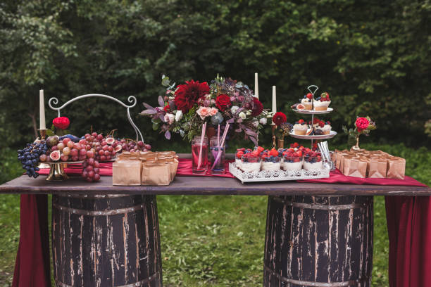 istock Wedding reception wooden table standing on two barrels, decorated with red flowers, sweets, drinks and silk. Rustic style 1026273382