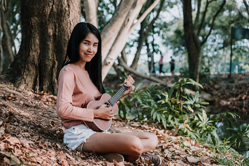 Happy woman smiling and playing ukulele under tree at park in summer.