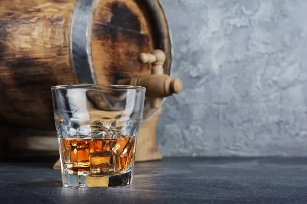 Strong alcoholic drink scotch whisky with ice cubes in old fashion glass and vintage wooden barrel in cellar on gray concrete background