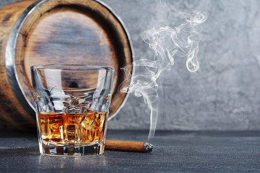 Strong alcoholic drink scotch whisky with ice cubes in old fashion glass with smoking cigar and vintage wooden barrel in cellar on gray concrete background