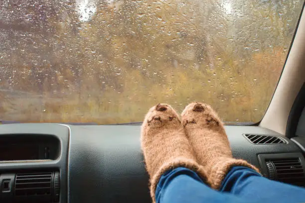 Woman legs in warm cute socks on car dashboard. Drinking warm tee on the way. Fall trip. Rain drops on windshield. Freedom travel concept. Autumn weekend