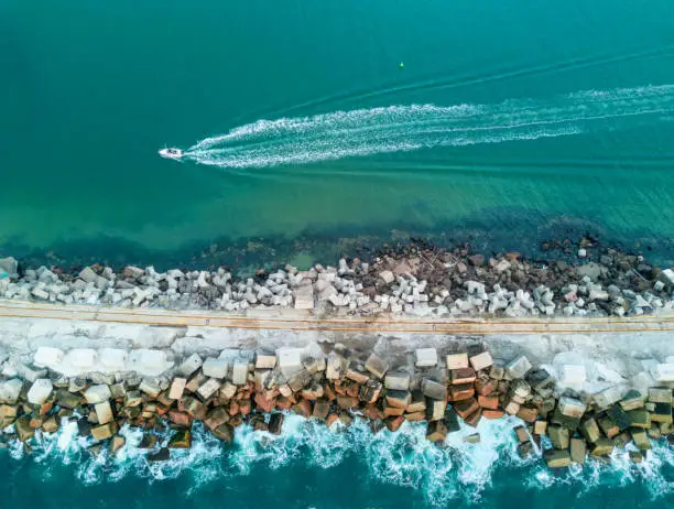 Aerial views of a speed boat leaves a wake behind it as it travels alongside a breadwall made up of concrete blocks and rough cut rock.