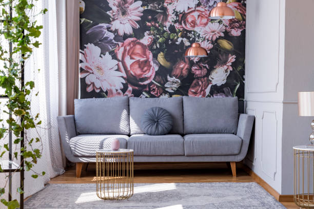sonnendurchflutete, grau sofa mit einer floralen print mauer im winkel eine feminine wohnzimmer interieur mit goldenen accessoires - kunstdruck fotos stock-fotos und bilder