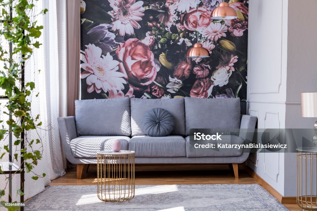 Sunlit, gray sofa by a floral print wall in the nook of a feminine living room interior with golden accessories Wallpaper - Decor Stock Photo