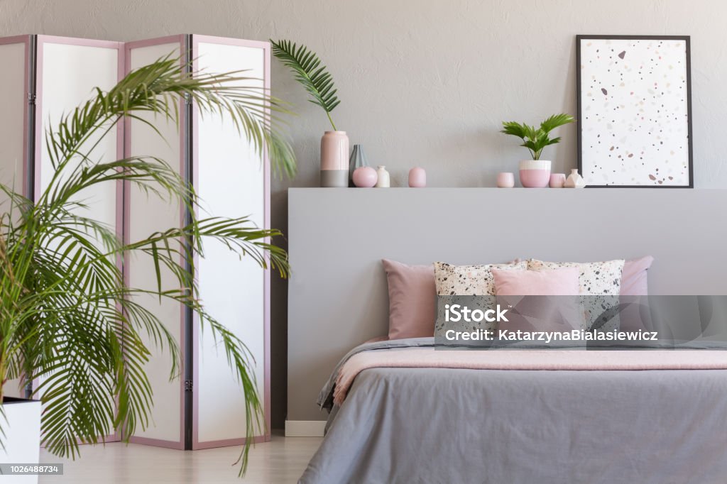 Pink pillows on grey bed in pastel bedroom interior with palm and poster on bedhead. Real photo Apartment Stock Photo