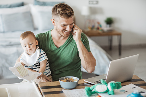 Father using mobile phone to finish some work from home and baby playing with some paper, they are at kitchen table