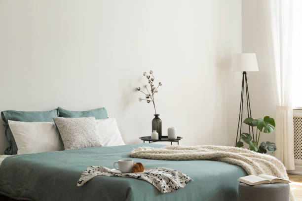 bedroom interior with sage green and white sheets and cushions and a blanket. black metal table with vases beside the bed. a lamp standing in the corner. real photo. - sheet metal fotos imagens e fotografias de stock