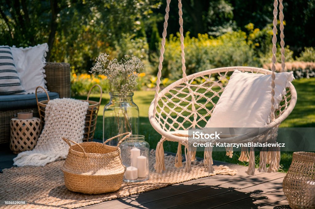 A beige string swing with a pillow on a patio. Wicker baskets, a rug and a blanket on a wooden deck in the garden. Patio Stock Photo