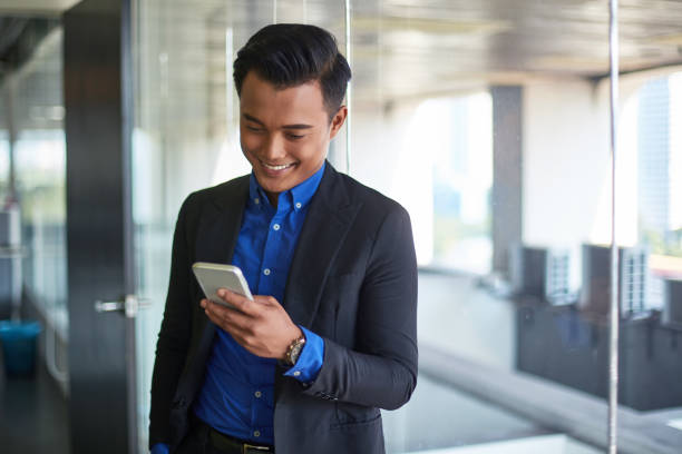 Smiling businessman using mobile phone in office Smiling young male professional using mobile phone. Confident businessman is text messaging on smart phone. He is wearing suit in office. malaysia office workers stock pictures, royalty-free photos & images