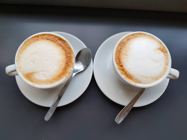 istock Loving coffee concept, Top view two white cup cappuccino or coffee cup latte on dark table near window with light shade on tabletop at cafe. 1026535086