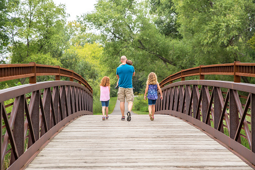 Rear view of a father and three children walking across a wooden and steel bridge in the woods on a late summer day. The father is carrying his toddler son in his arms while his daughters walk on both sides of him.