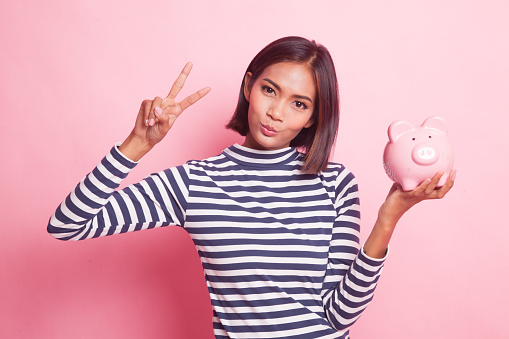 Asian woman show victory sign with pig coin bank on pink background