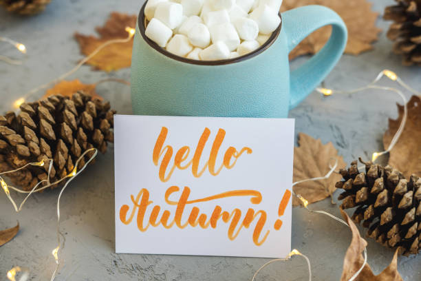 Blue mug with coffee, hot chocolate or cocoa with marshmallow on gray concrete background and lying dry leaves, cones, garland, sign Hello Autumn. Concept of warm cozy autumn.