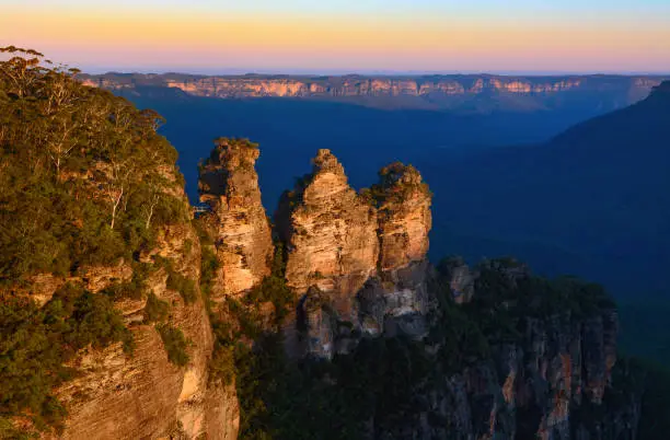 Evening sunlight highlights the peaks of the Three Sisters rock formation against the shadowed Jamison Valley in the background just before sunset in Australia's beautiful Blue Mountains.