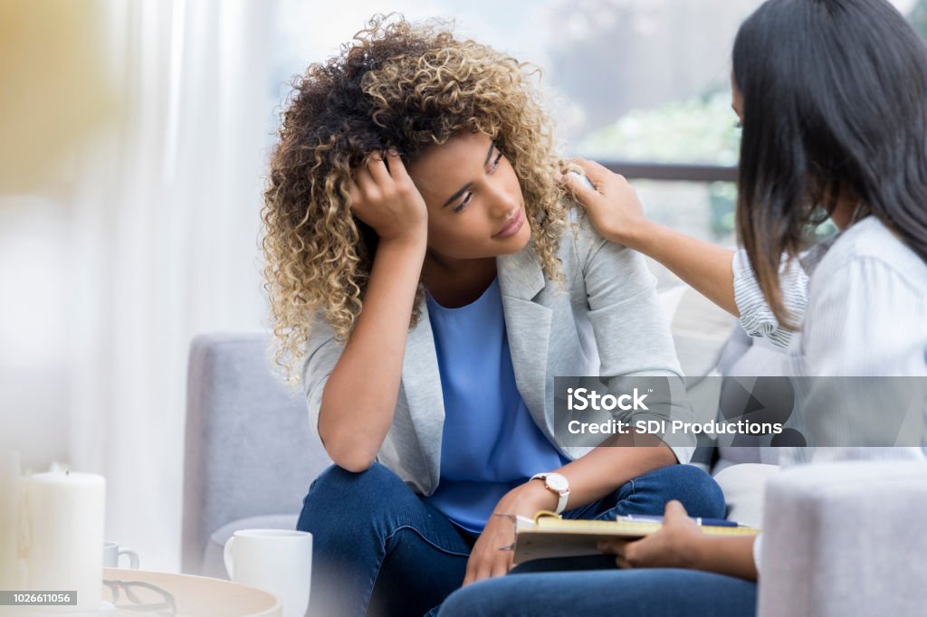 Depressed young woman talks to therapist A young woman sits on a couch with her unrecognizable therapist.  She puts her head in her hand as she looks out the window with a sad expression.  Her therapist puts a hand on her shoulder. Mental Health Stock Photo