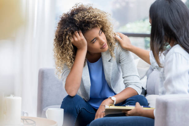 Depressed young woman talks to therapist A young woman sits on a couch with her unrecognizable therapist.  She puts her head in her hand as she looks out the window with a sad expression.  Her therapist puts a hand on her shoulder. overworked photos stock pictures, royalty-free photos & images