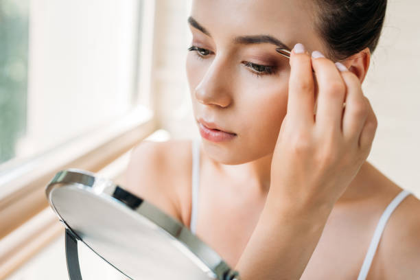 istock young woman correcting eyebrows with tweezers and looking at mirror 1026612140