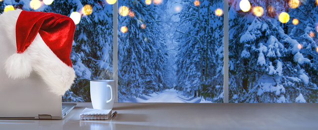 Laptop on table with Santa Claus hat at home with panoramic view through window of snowy trees in winter forest