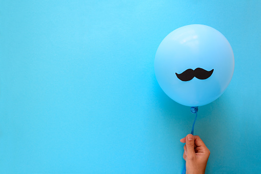 Hand holding blue balloon with a paper mustache on blue paper background. Cut out style. Movember  or man health concept. Top view. Flat lay. Copy space