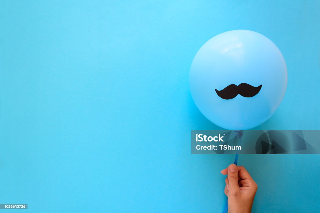 Hand holding blue balloon with a paper mustache on blue paper background. Cut out style. Movember  or man health concept. Top view. Flat lay. Copy space Men Stock Photo