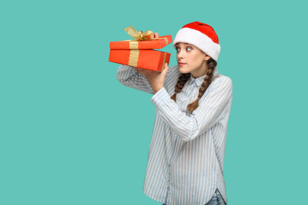 New year concept. cunning beautiful girl in striped light blue shirt in red christmas cap standing holding red gift box, unboxing and looking inside. New year concept. cunning beautiful girl in striped light blue shirt in red christmas cap standing holding red gift box, unboxing and looking inside. indoor, studio shot isolated on green background. christmas excitement stock pictures, royalty-free photos & images